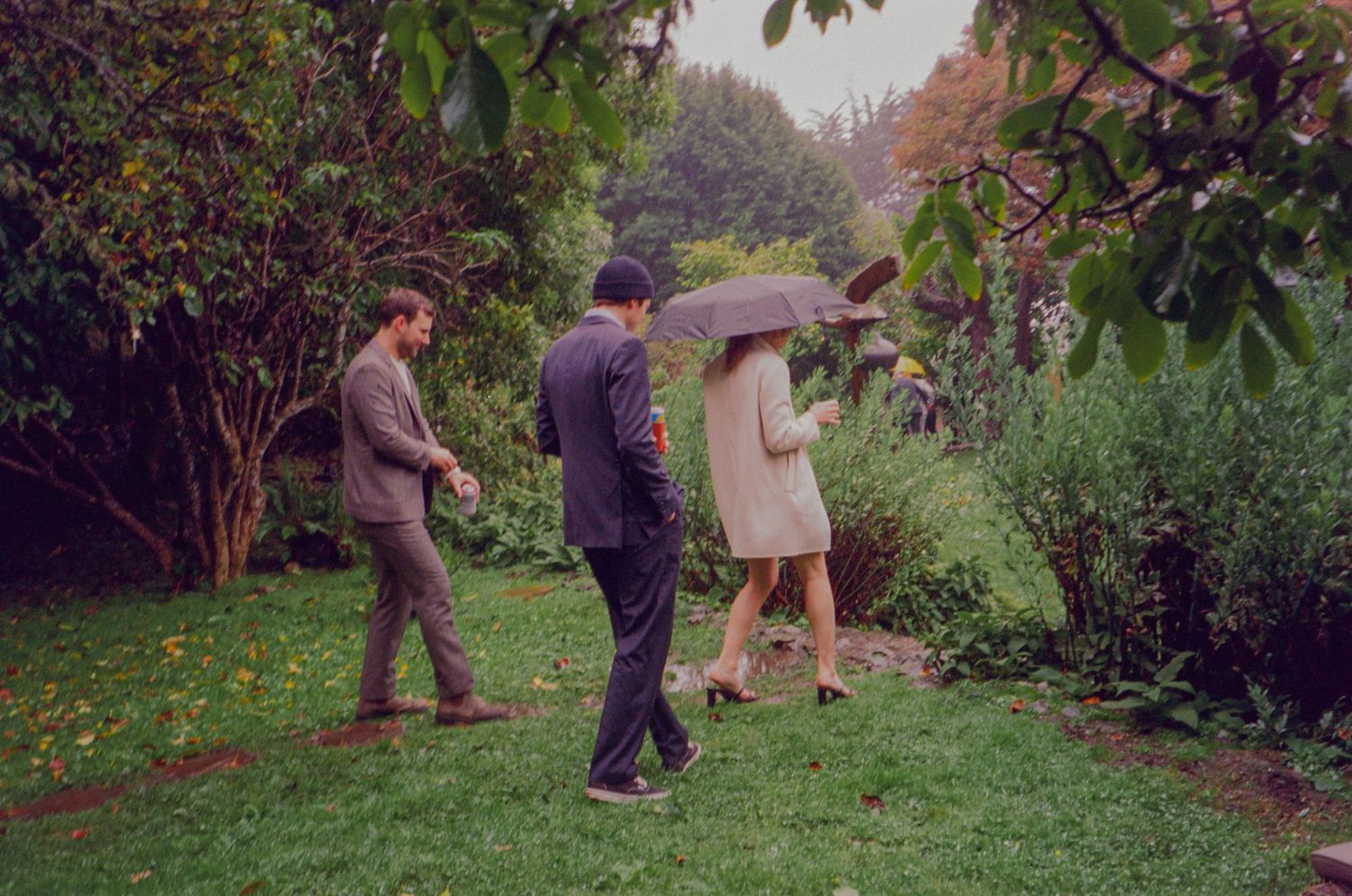 Three people walk through a lush garden in the rain, one sheltering under an umbrella.