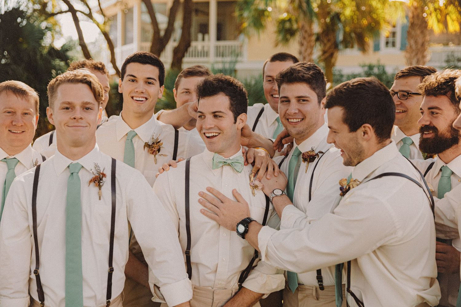 Groomsmen in white shirts with mint ties and suspenders laughing together at an outdoor wedding.