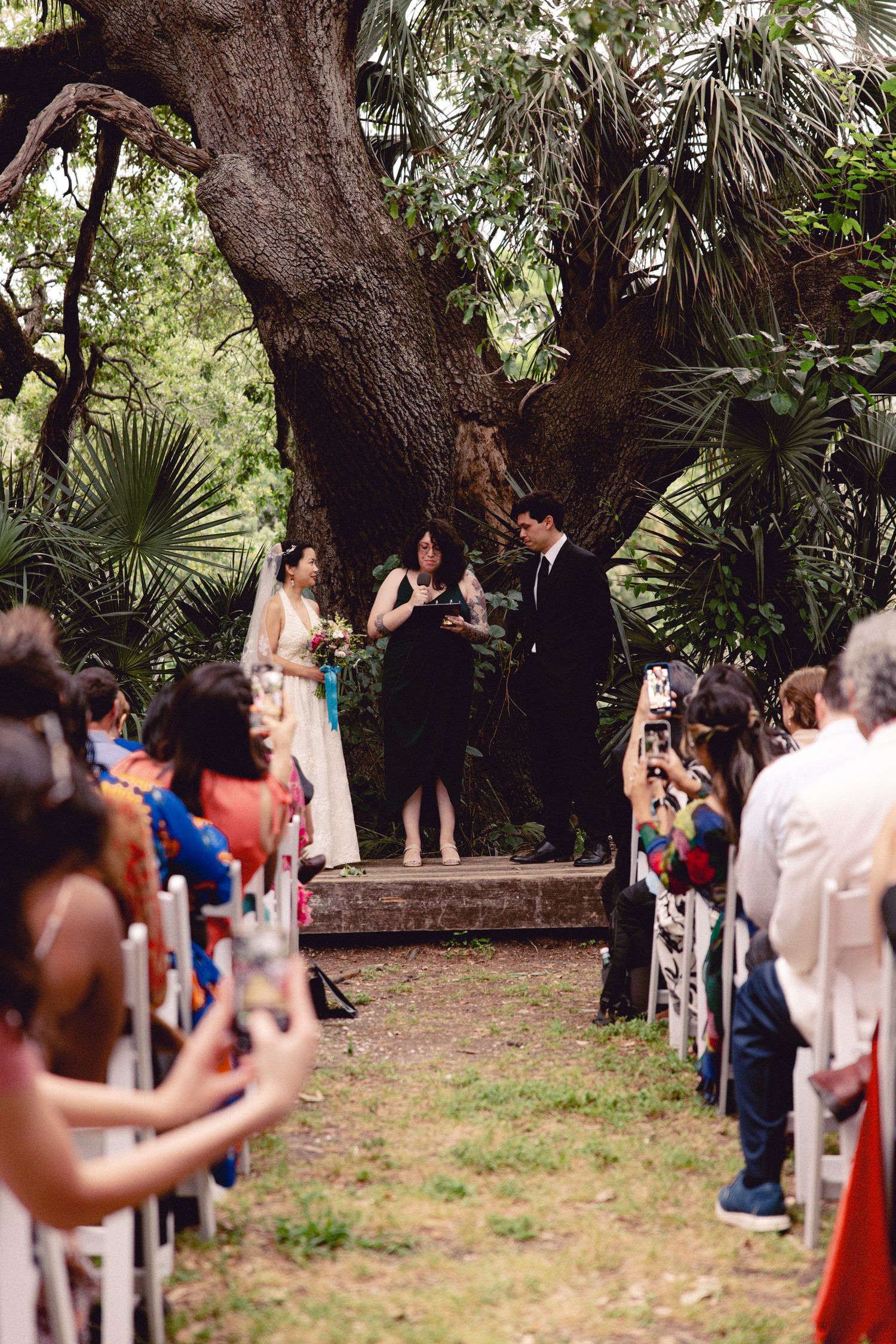 Wide aisle view of outdoor wedding ceremony with guests seated in white chairs amid tropical trees.