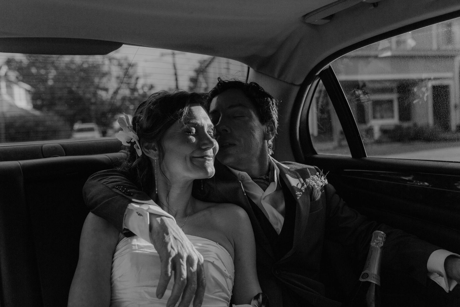 Black and white photo of newlyweds relaxing together in vintage car with champagne after wedding.