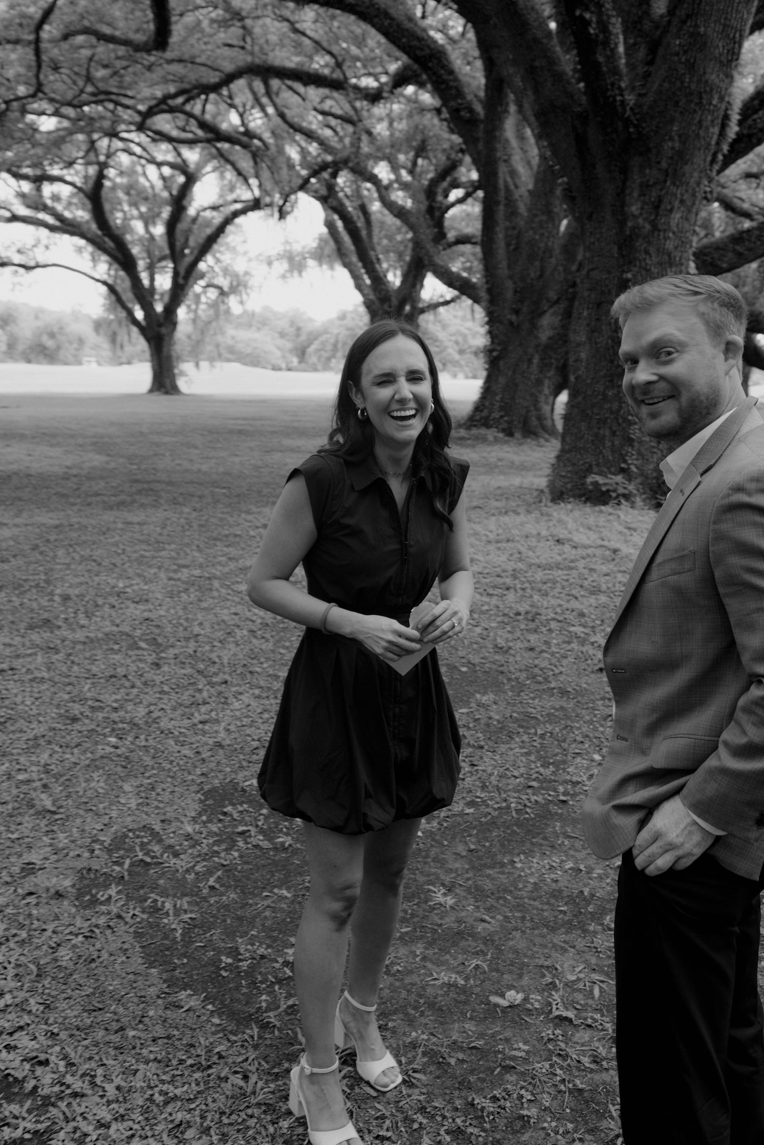 Black and white photo of a laughing woman in a dress and a man in a suit standing outdoors under large oak trees.