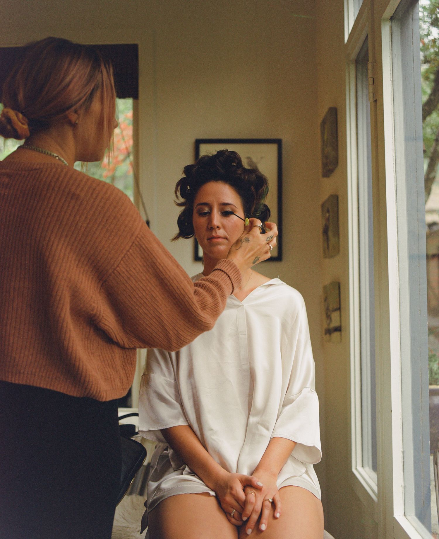 Makeup artist applying eye makeup to a bride seated by a bright window during wedding preparations.