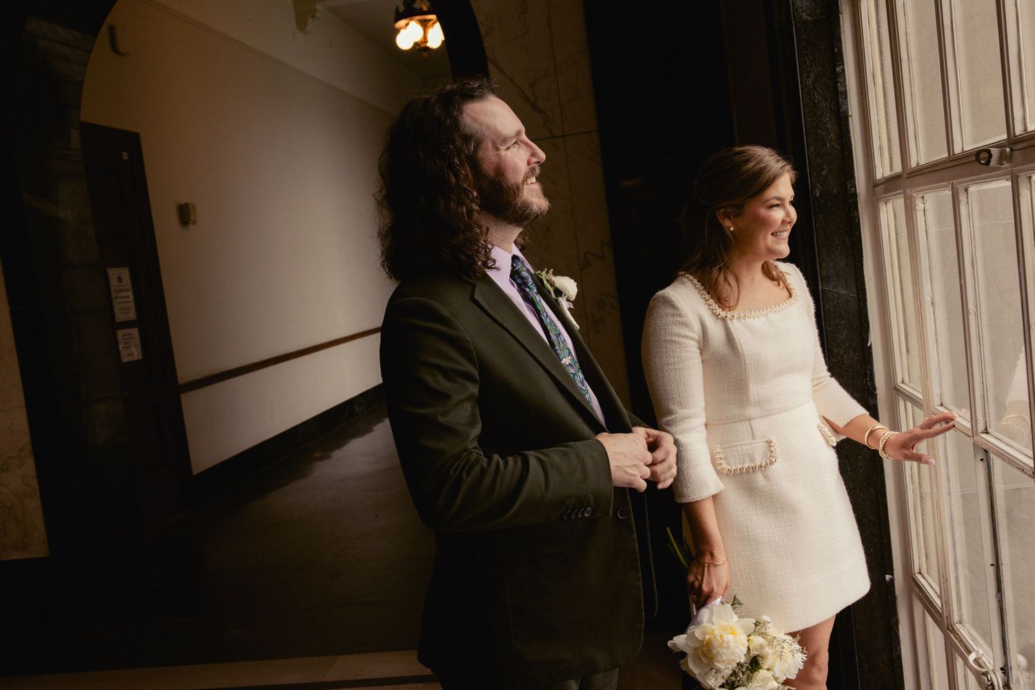 Bride in short white dress and groom in dark suit stand by window in dimly lit venue.