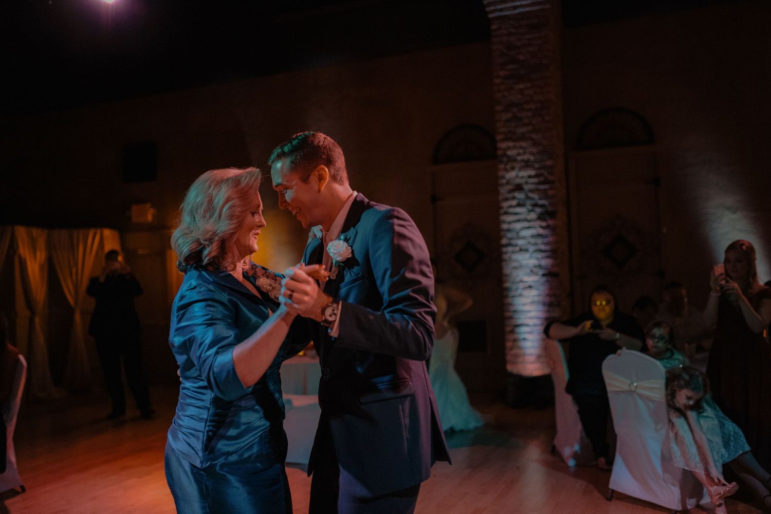 Groom shares a warm mother-son dance under amber lighting at elegant wedding reception.
