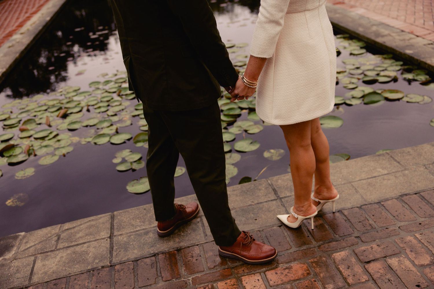 Couple holding hands in matching white mini dress and dark suit walking past a lily pad pond.