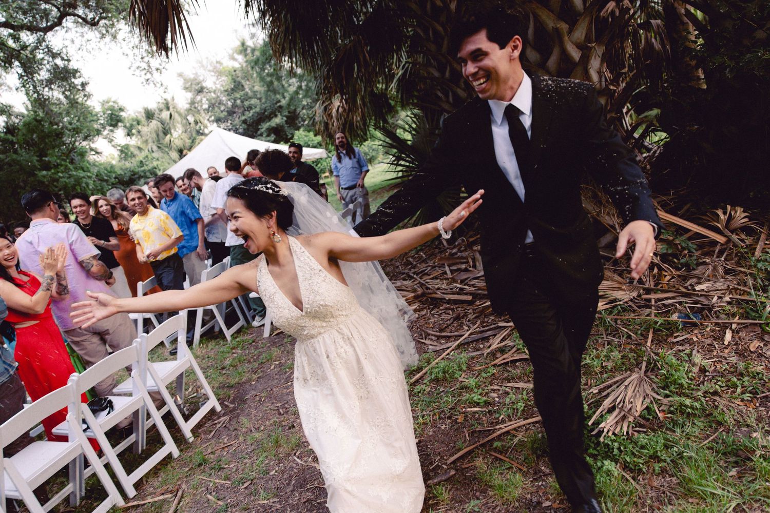 Laughing bride and groom exit outdoor wedding ceremony between rows of white chairs and cheering guests.