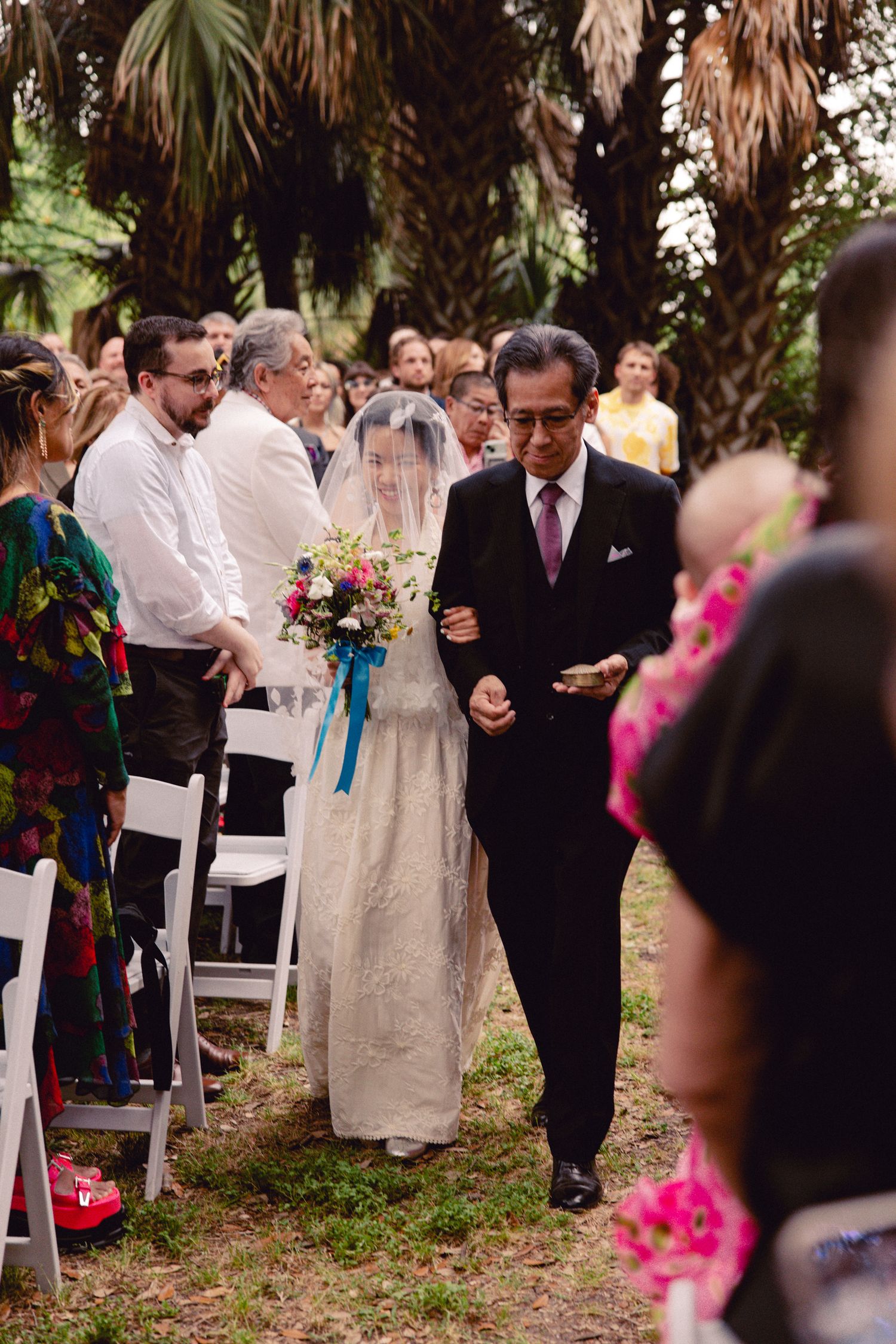 Father walks bride down the aisle in a lush outdoor garden wedding ceremony with colorful guests.