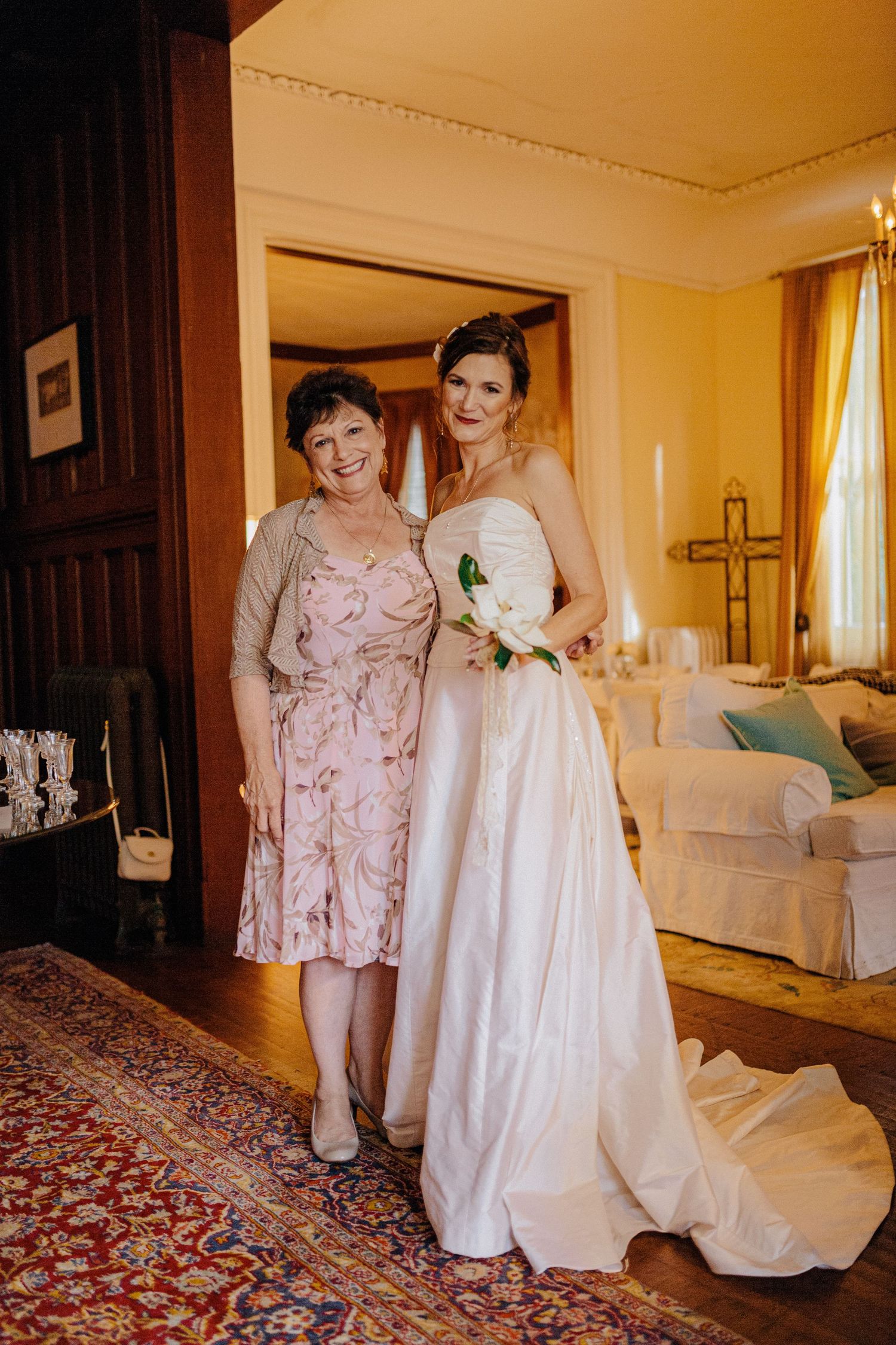 Bride in strapless white gown poses with smiling mother in pink dress inside elegant home.