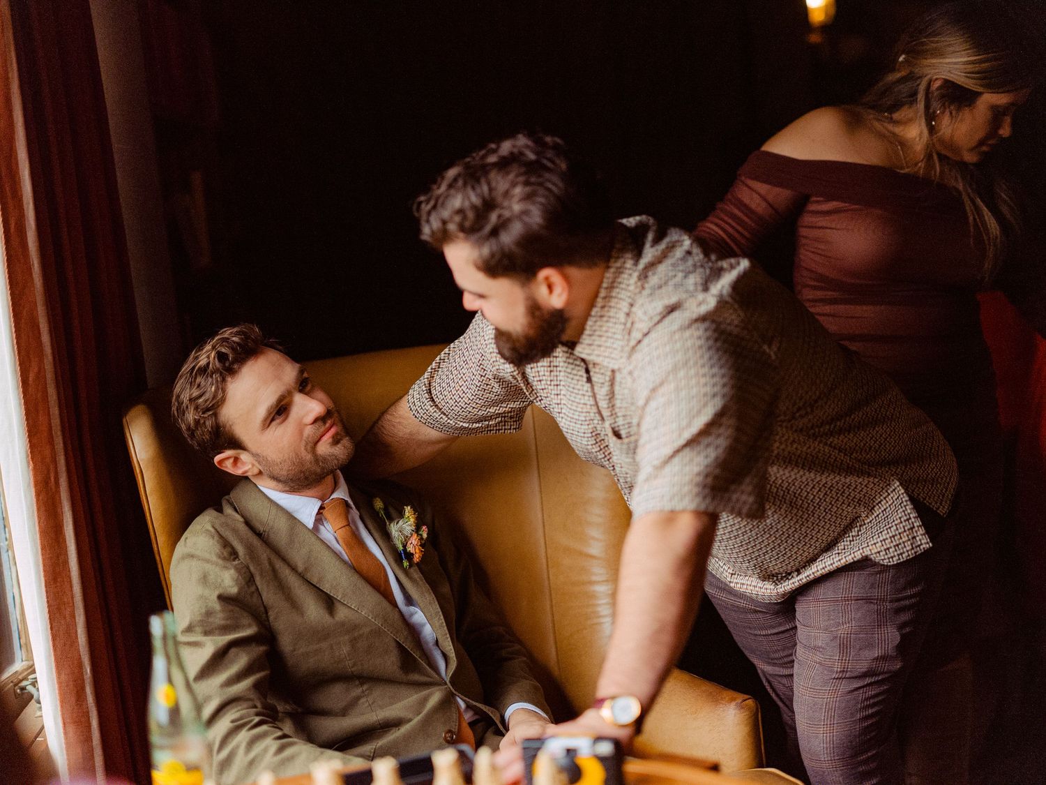 Groom in tweed suit relaxes in leather chair as friend touches his face before wedding ceremony.