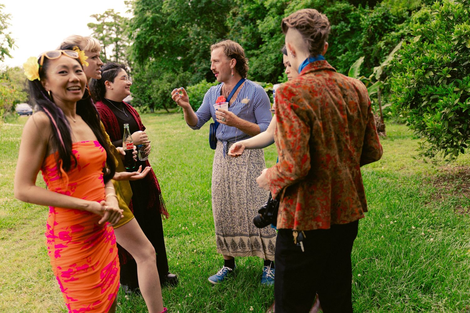 Colorfully dressed wedding guests laugh and socialize on a green lawn at an outdoor celebration.