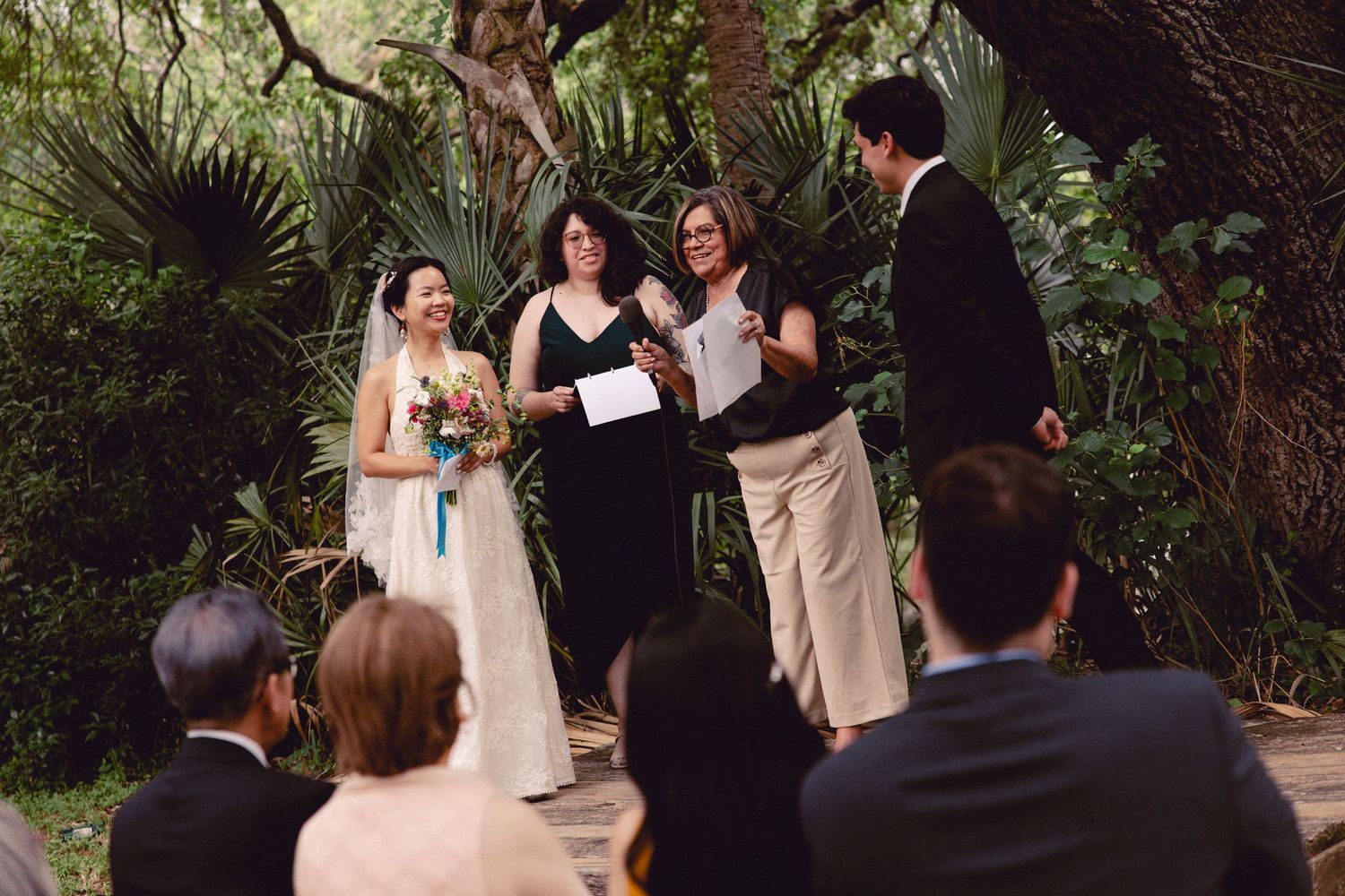 Bride and groom stand with officiant and witness during intimate outdoor wedding ceremony in garden.