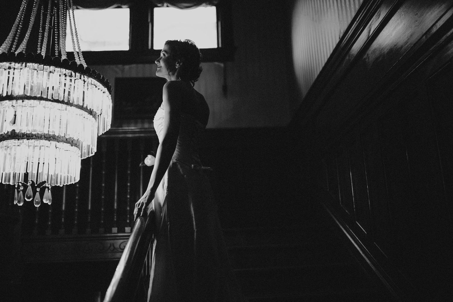 Elegant black and white silhouette of a bride standing beneath a crystal chandelier on a staircase.