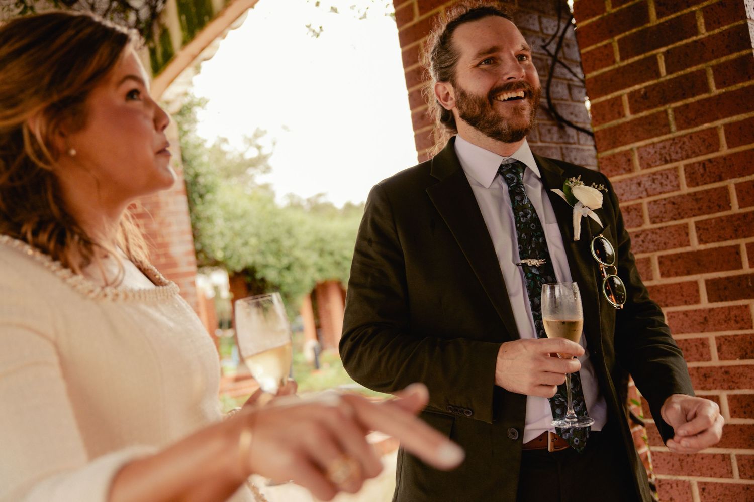 Bearded groom in dark suit laughing with bride at outdoor wedding reception.