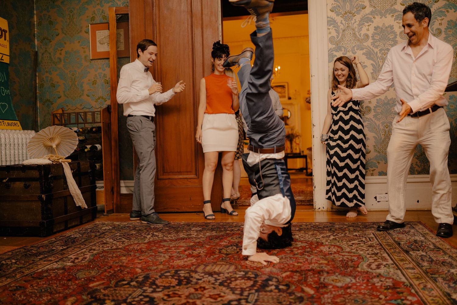 Wedding guest performing a headstand on a rug while guests cheer and laugh around him.