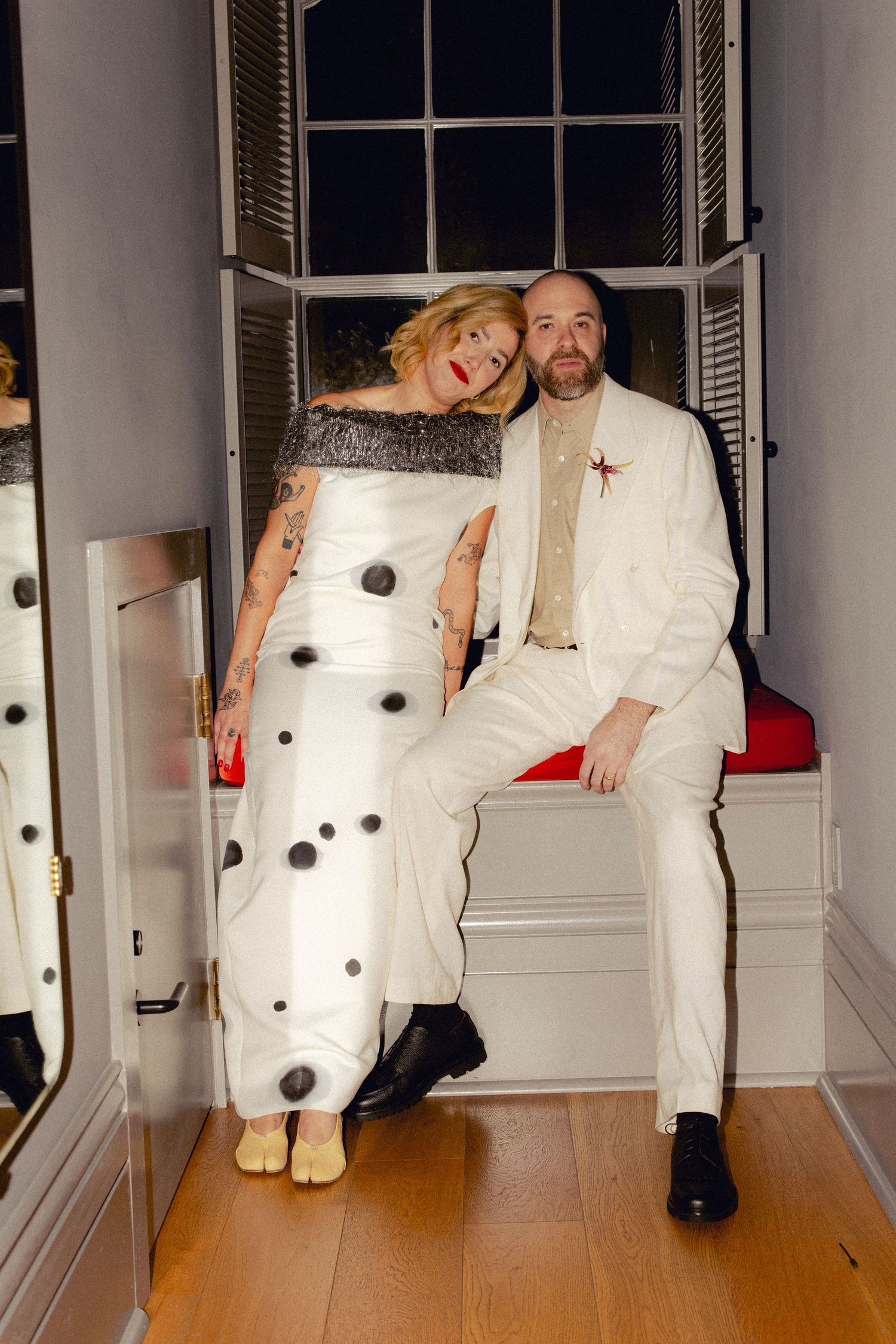 Smiling couple in eclectic wedding attire pose together at window seat during wedding reception.
