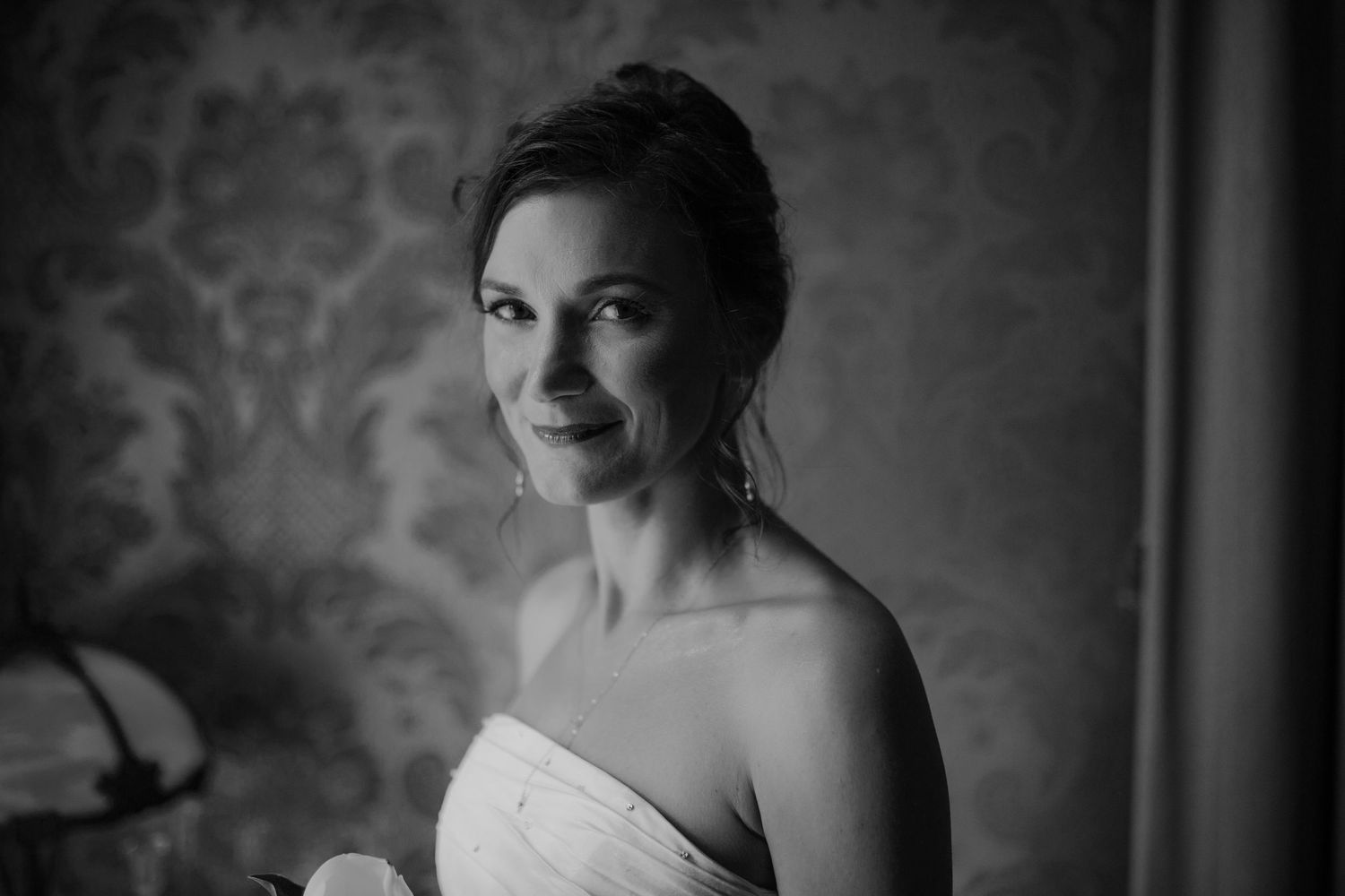Elegant bride in strapless gown smiling softly in black and white portrait with ornate wallpaper backdrop.