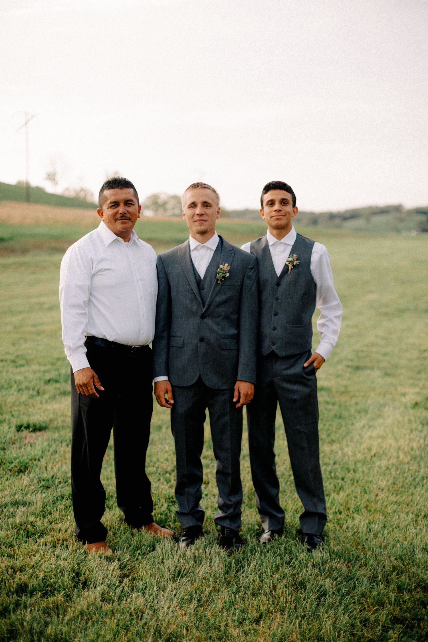 Groom in gray three-piece suit poses with two groomsmen on a green field at sunset.