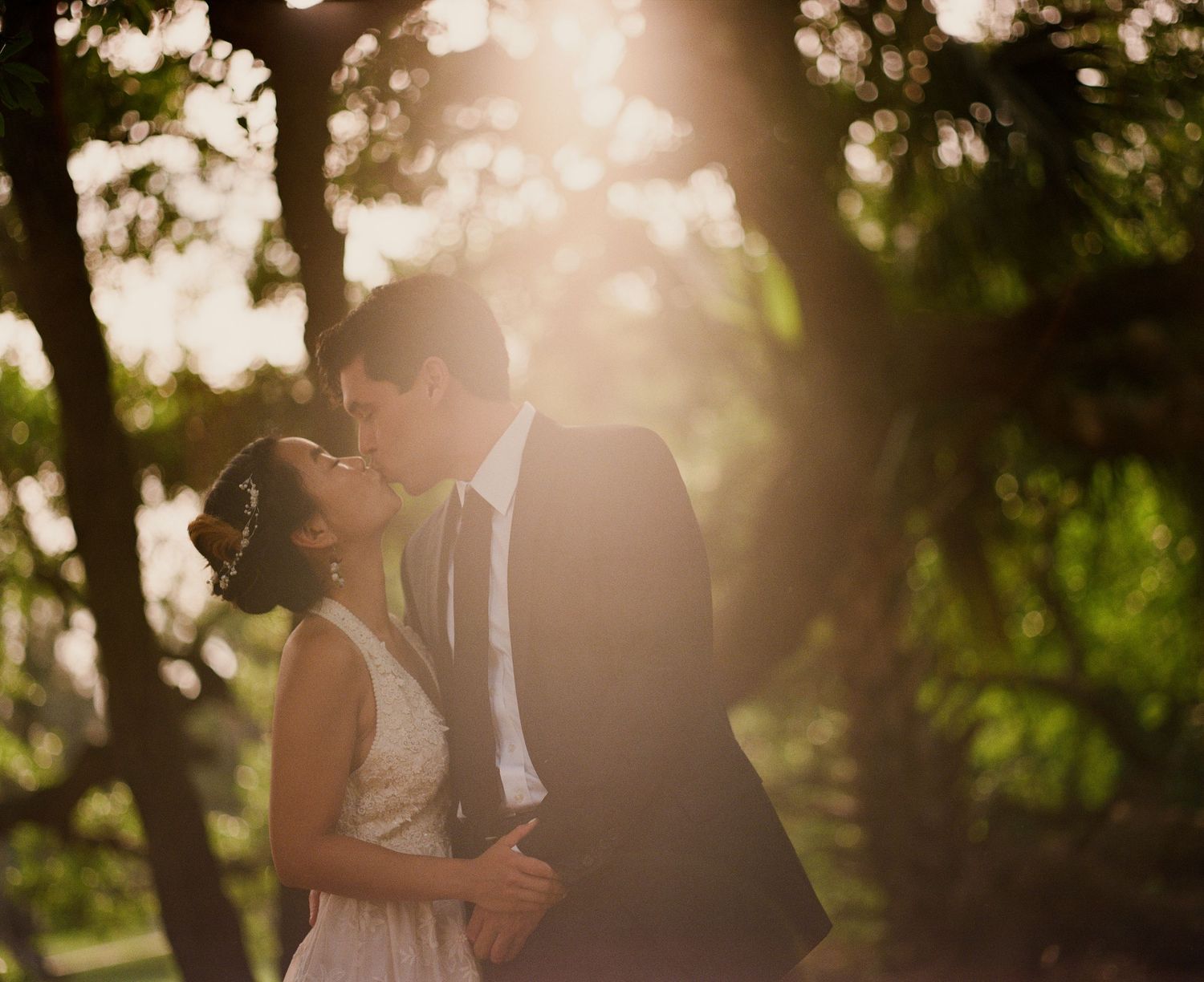 Bride and groom sharing a romantic kiss surrounded by golden sunlight filtering through trees.