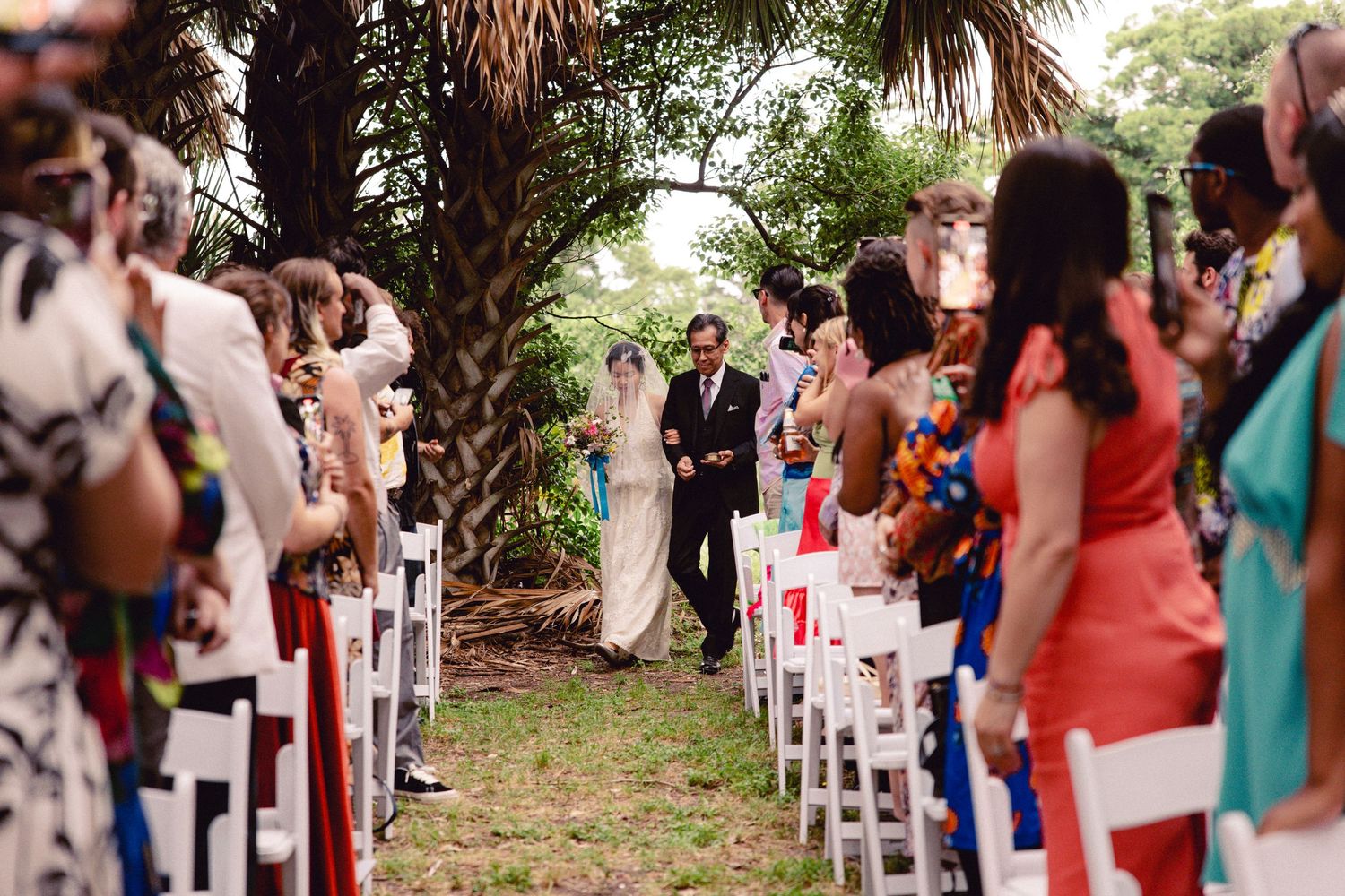 Father escorts bride in white gown down a long outdoor aisle lined with guests in colorful attire.