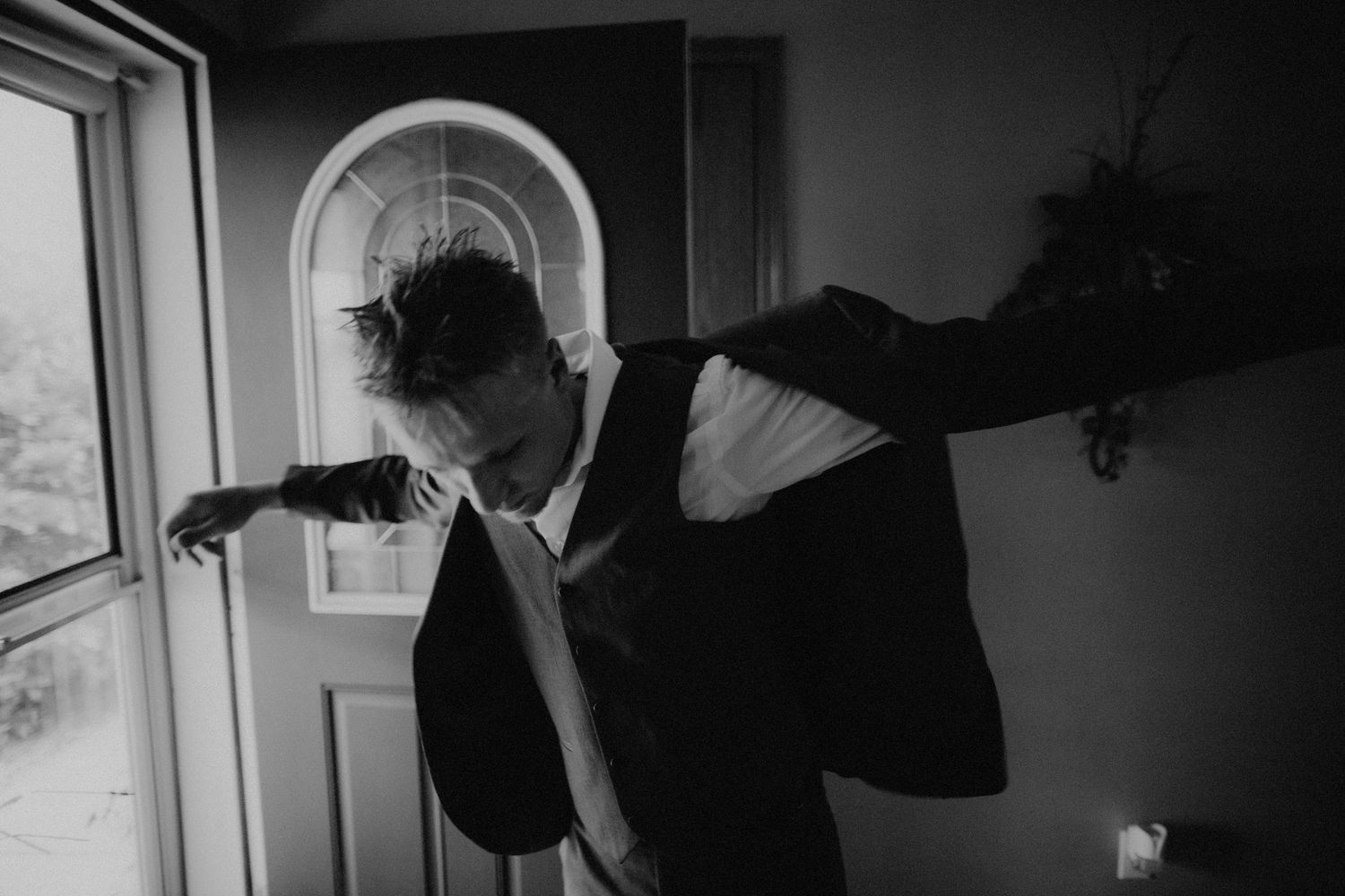 Groom putting on suit jacket near arched window in black and white photo.
