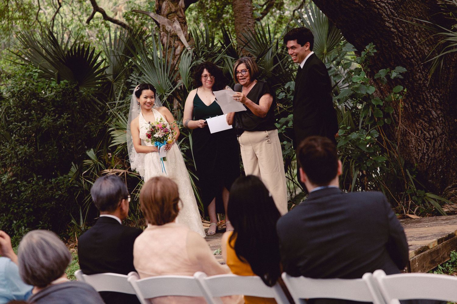 Wedding ceremony in a tropical garden setting with bride, groom, and officiant on a wooden platform.