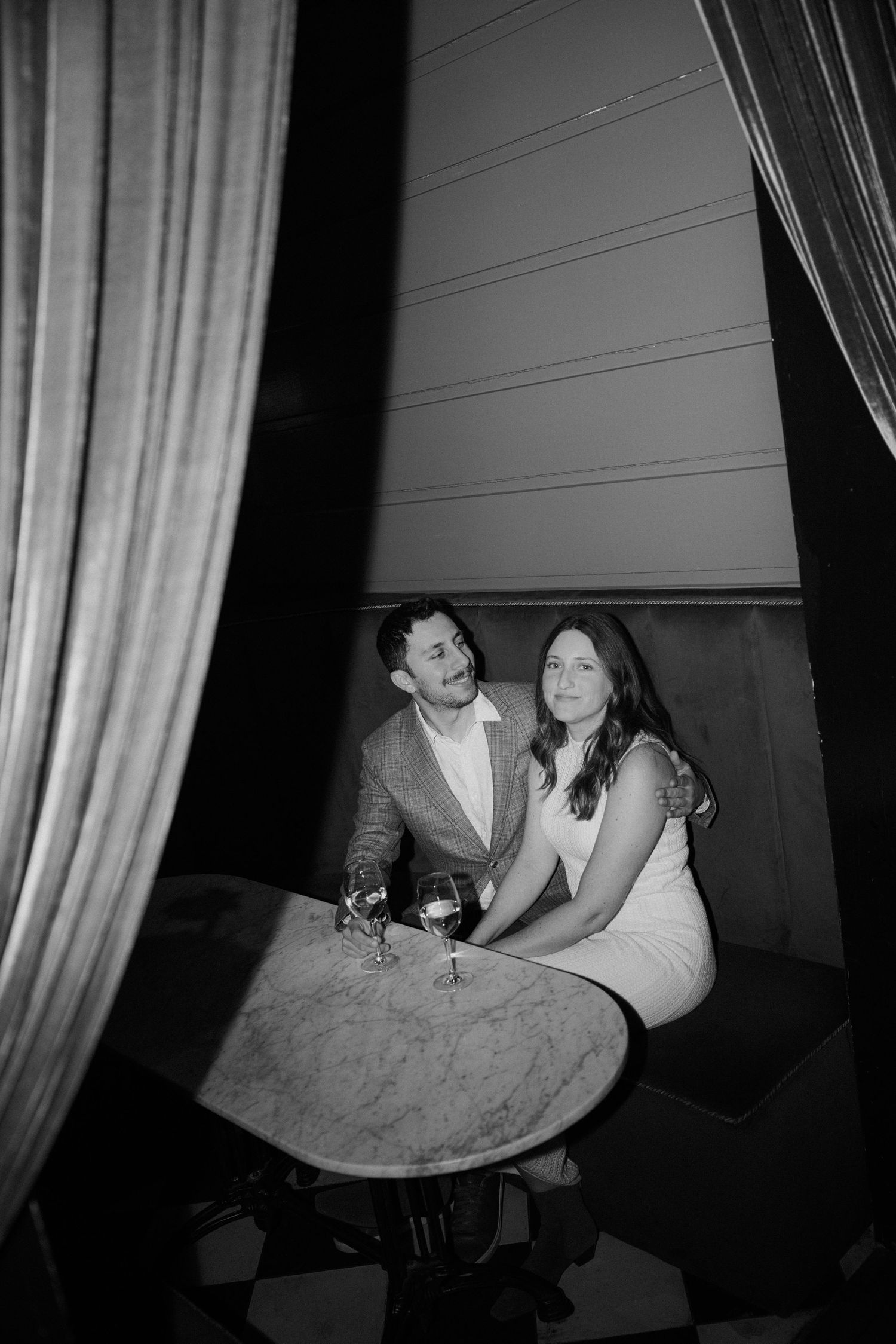 Couple sits intimately at a marble table with wine glasses in a black and white engagement photo.