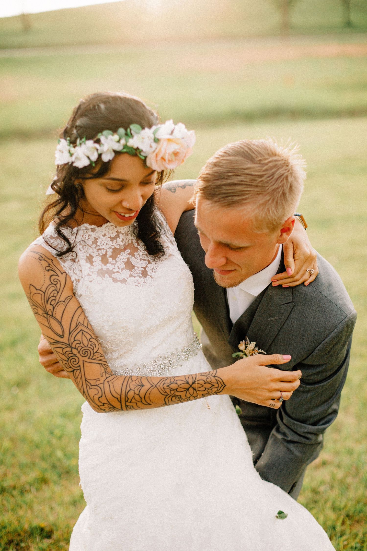 Bride with floral crown and tattoos embraces groom in gray suit during golden hour outdoor wedding.