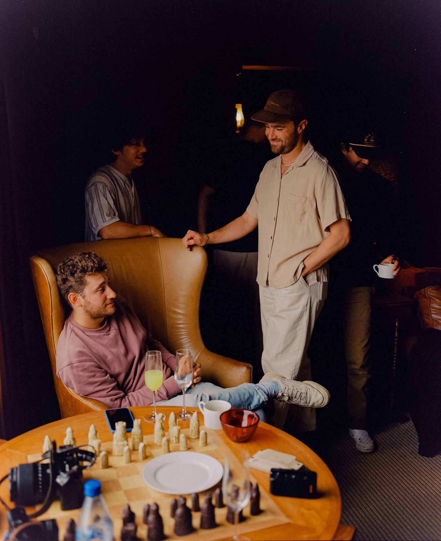 Men socializing around a chess set on a round table in a dimly lit lounge setting.
