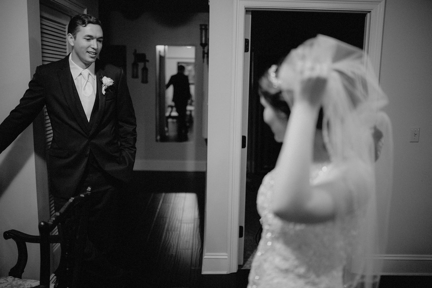 Groom in black suit watches bride adjusting her veil in a black and white wedding photo.