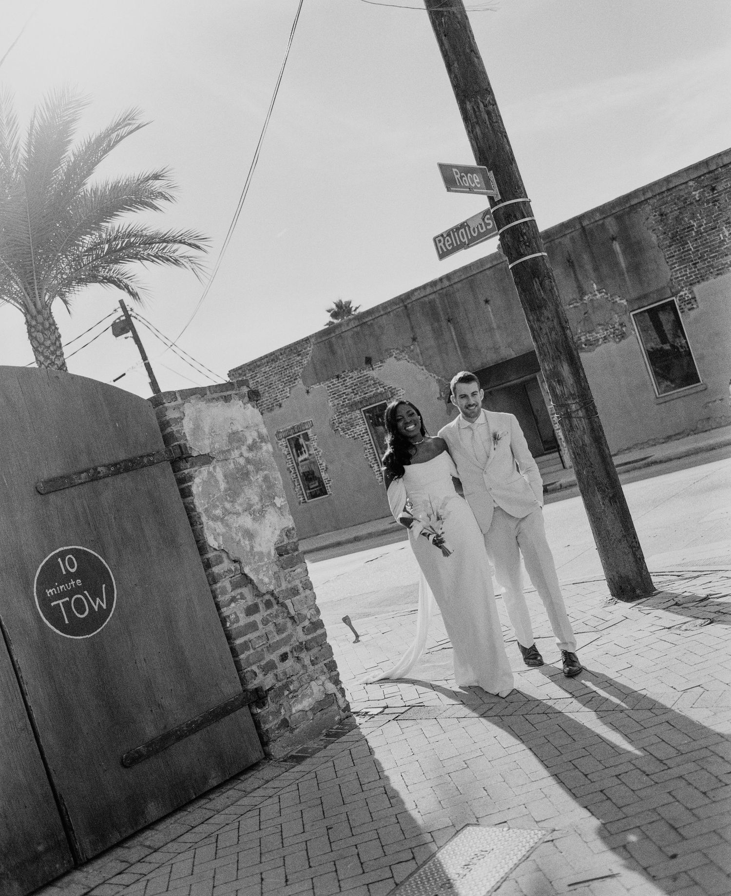 Bride and groom in white attire posing on sunny street corner with palm tree in black and white.