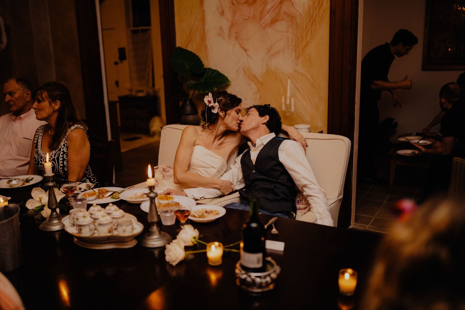 Bride and groom sharing romantic kiss at candlelit wedding reception dinner table with guests nearby.
