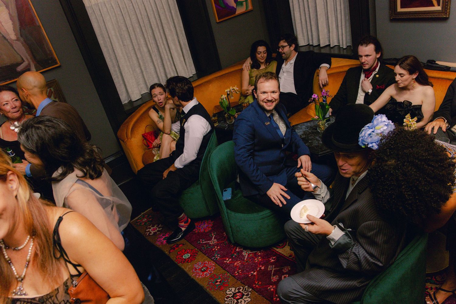 Group of elegantly dressed guests socializing on green velvet chairs at an upscale party.