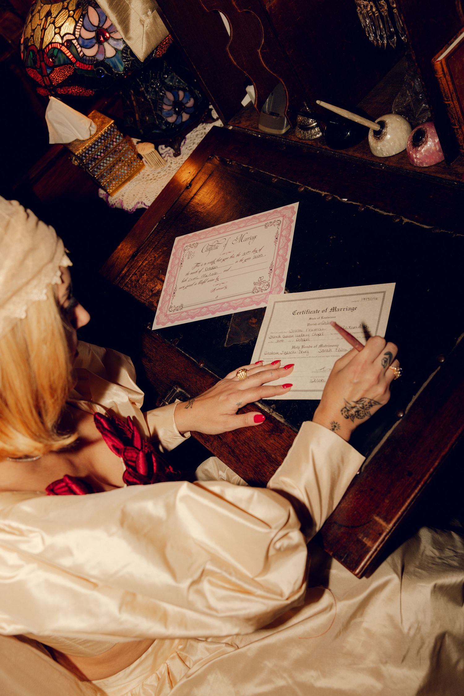 Bride with red nails and tattoos signing a marriage certificate at an antique wooden desk.