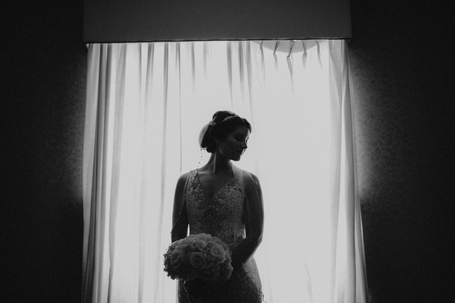 Silhouette of bride in lace wedding dress holding bouquet standing before sheer curtained window.