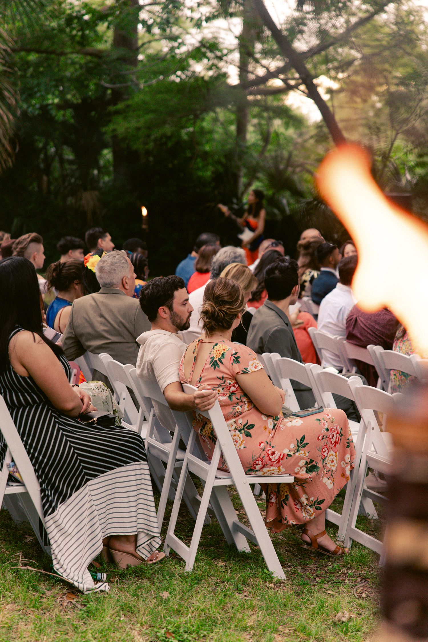 Wedding guests seated in white chairs at an outdoor garden ceremony at sunset with torchlight.