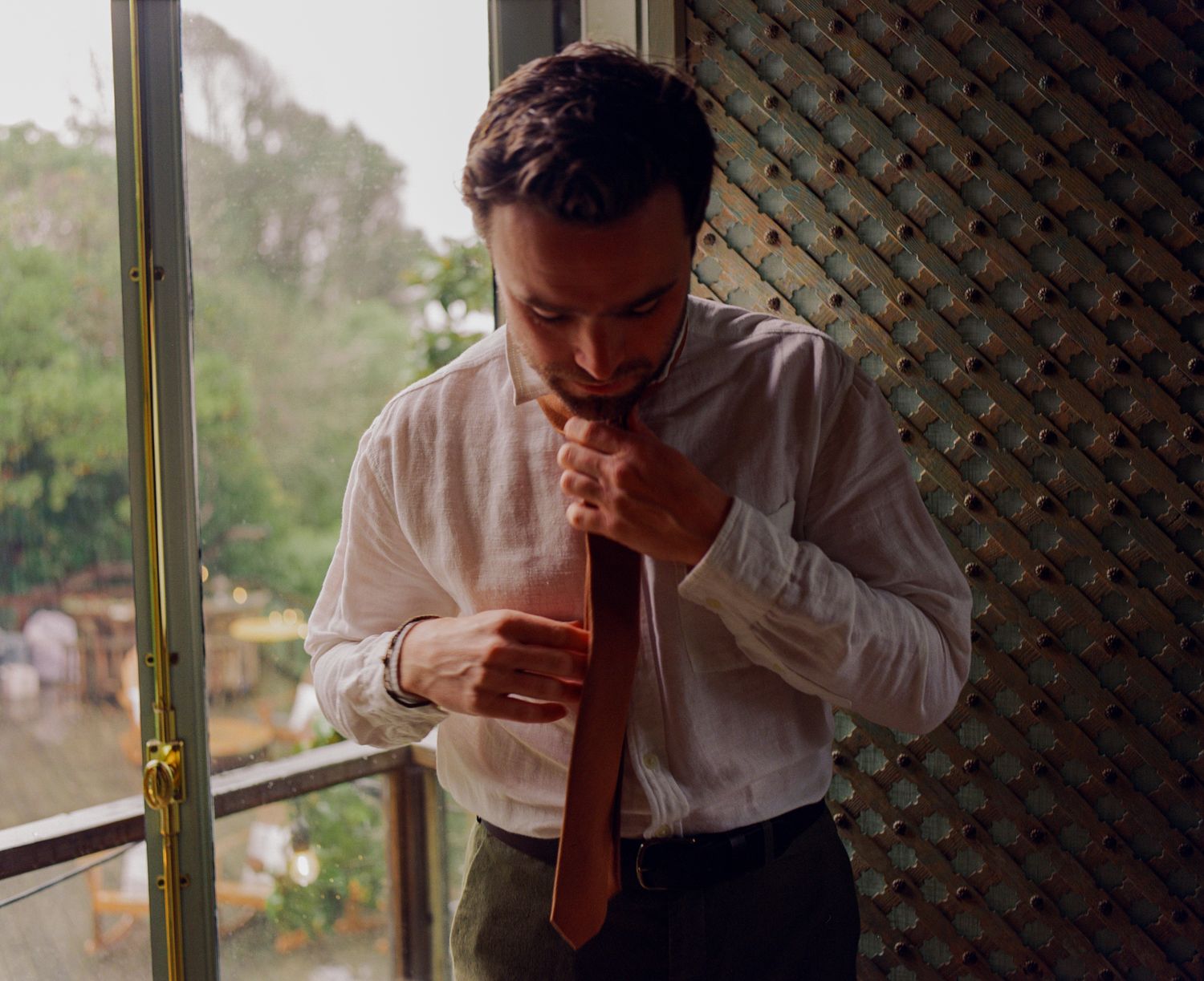 Groom tying his tie by a window with lush greenery in soft color wedding preparation photo.