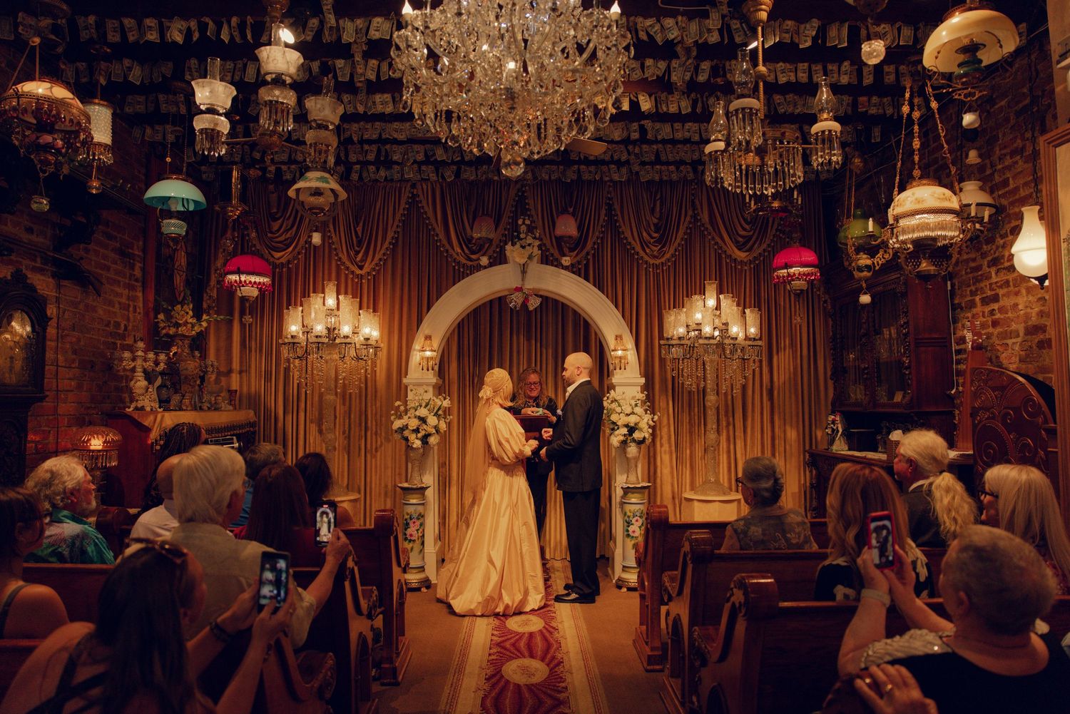 Wide-angle wedding ceremony shot under ornate arch surrounded by chandeliers and guests.
