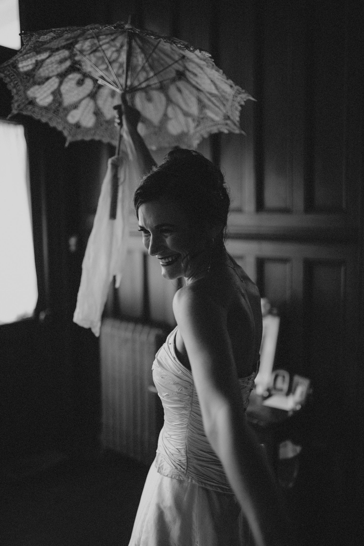 Black and white portrait of a smiling bride holding a lace parasol in a dimly lit room.