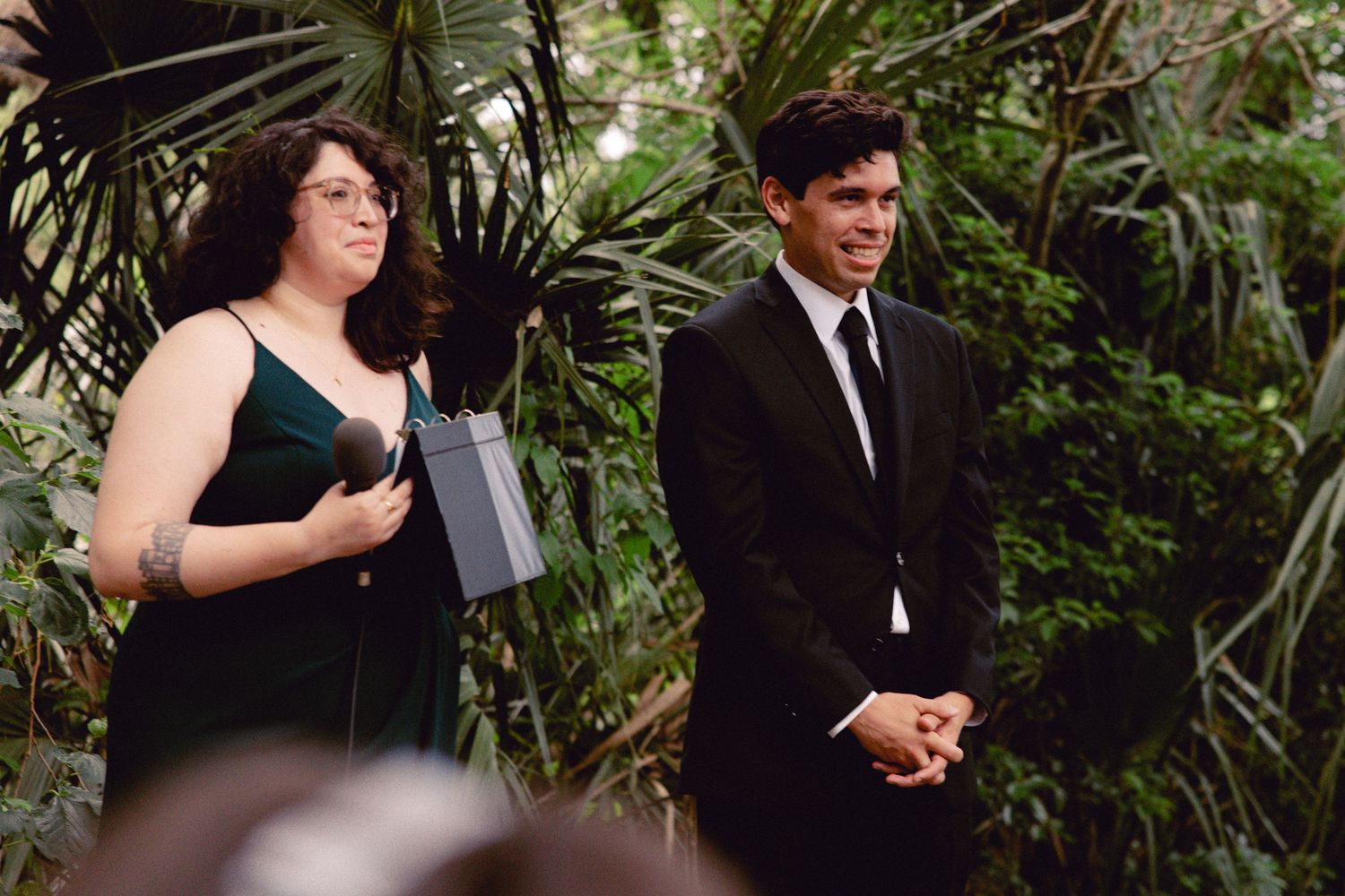 Groom in black tuxedo stands with officiant holding a book at an outdoor garden wedding ceremony.