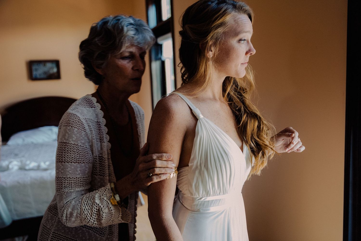 Mother helping bride into elegant white wedding gown in softly lit room.