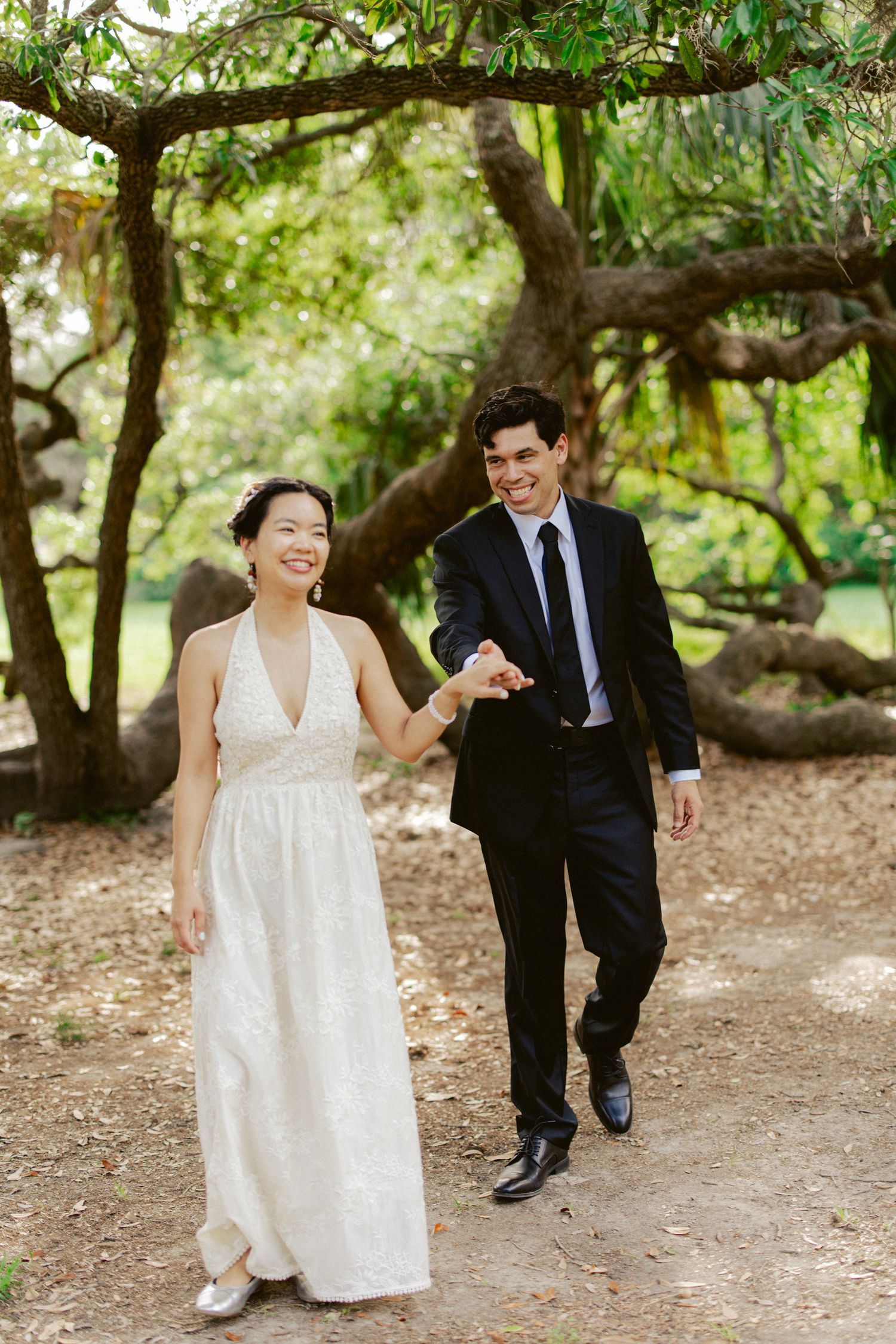Smiling bride in white halter gown and groom in black suit holding hands walking through lush garden.