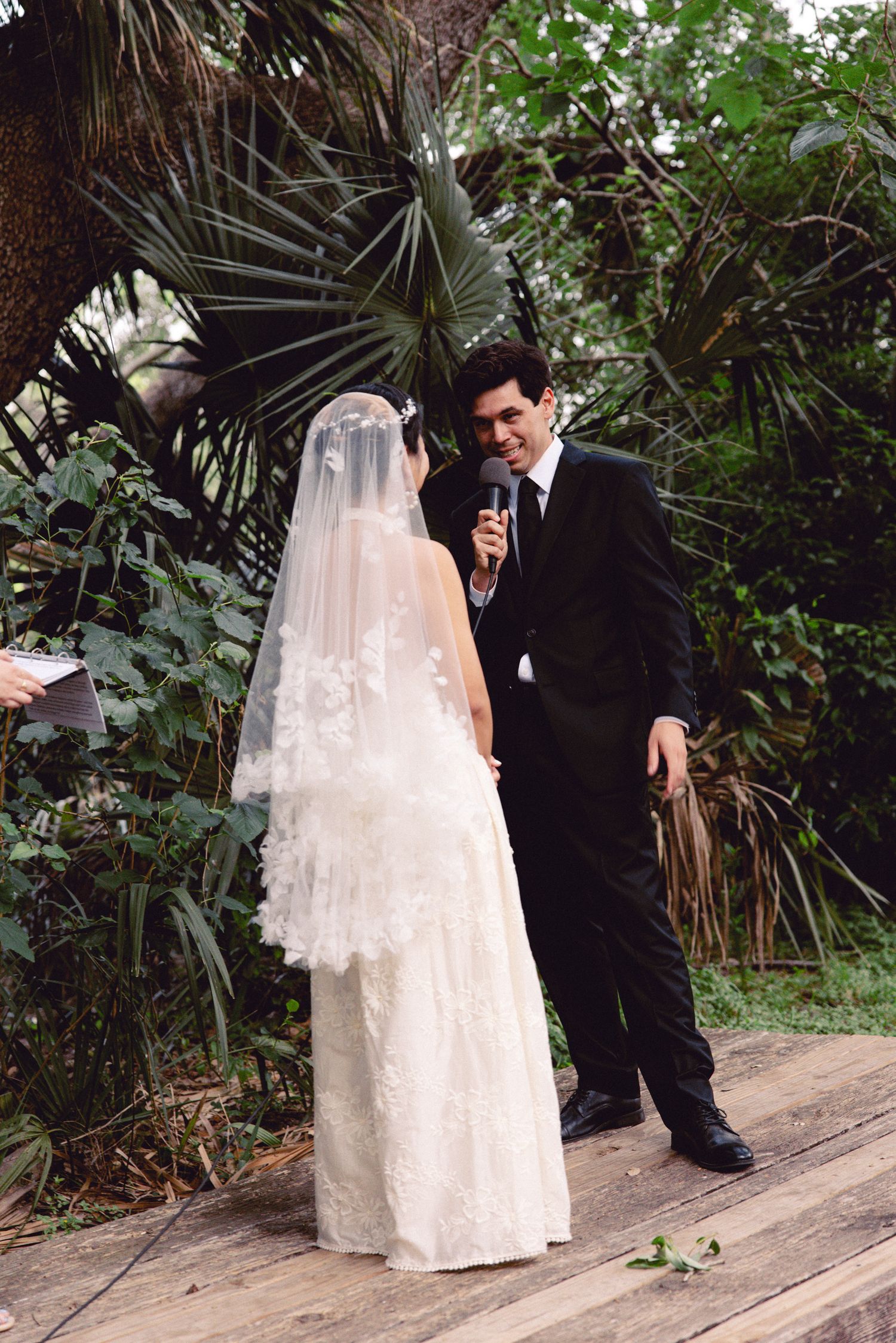Groom in black suit reads vows into microphone to veiled bride in lace wedding dress outdoors.