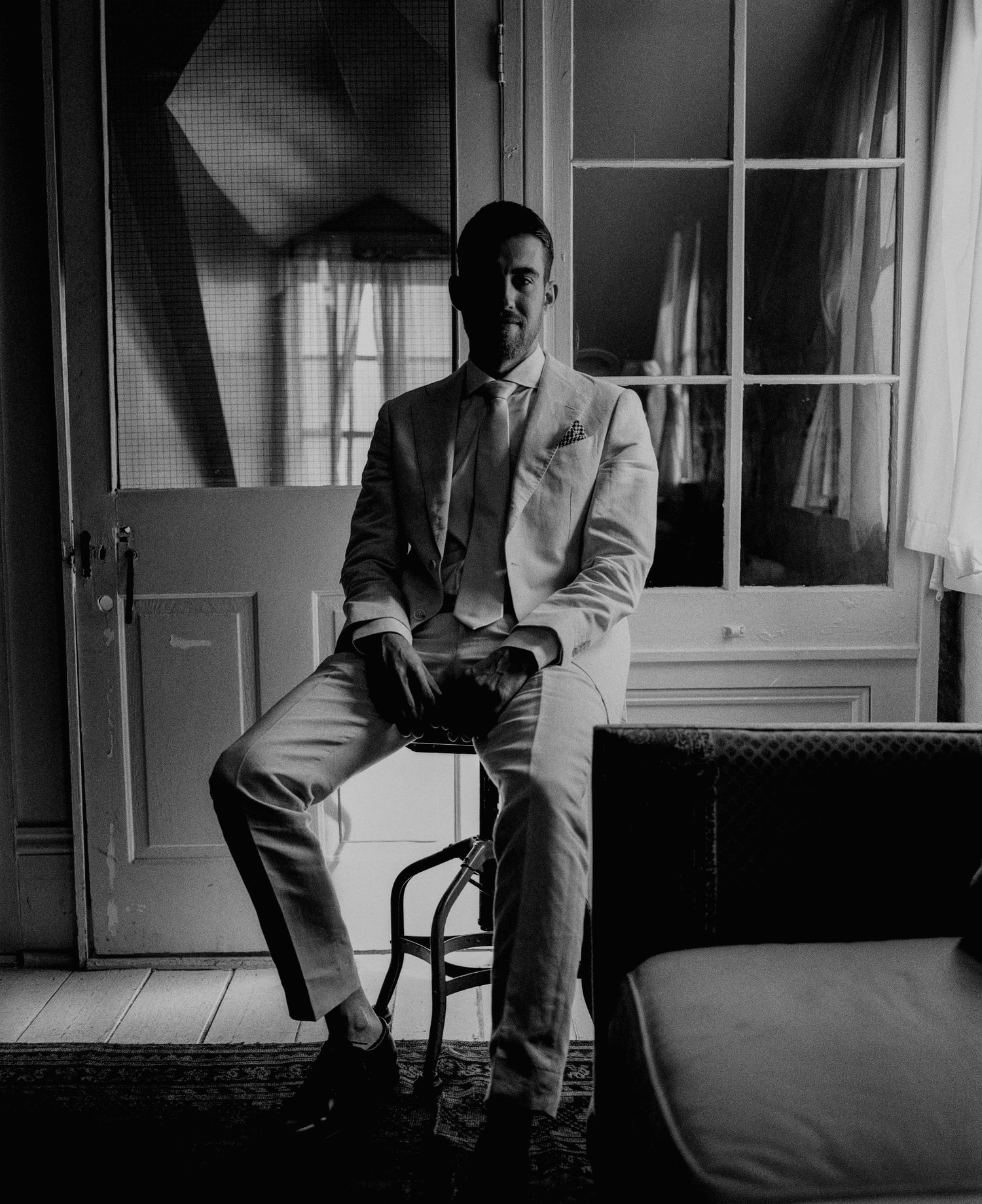 Groom in white suit sitting on stool before French doors in elegant black and white portrait.