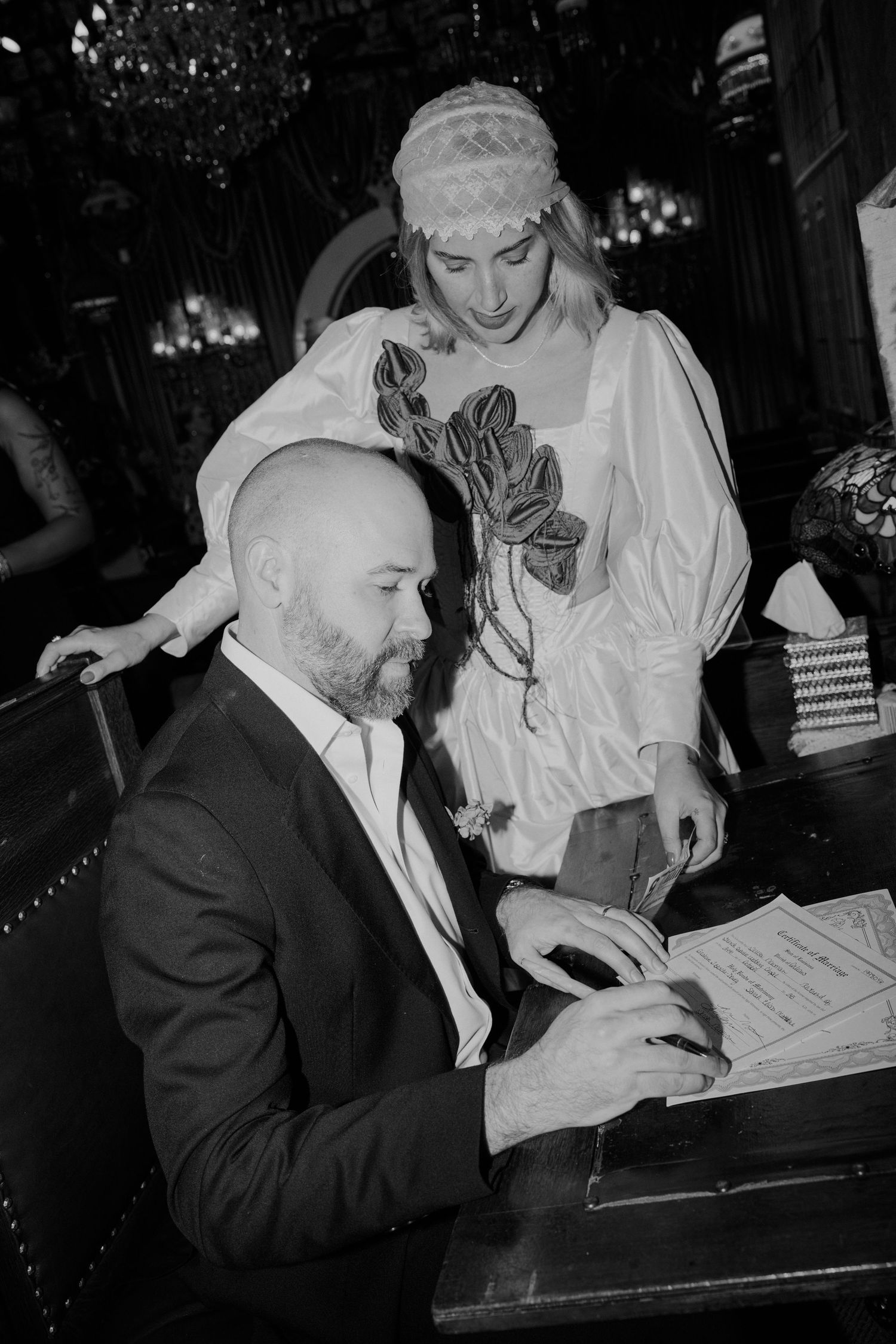 Bald groom in dark suit signs wedding documents as bride in white dress and headpiece looks on.
