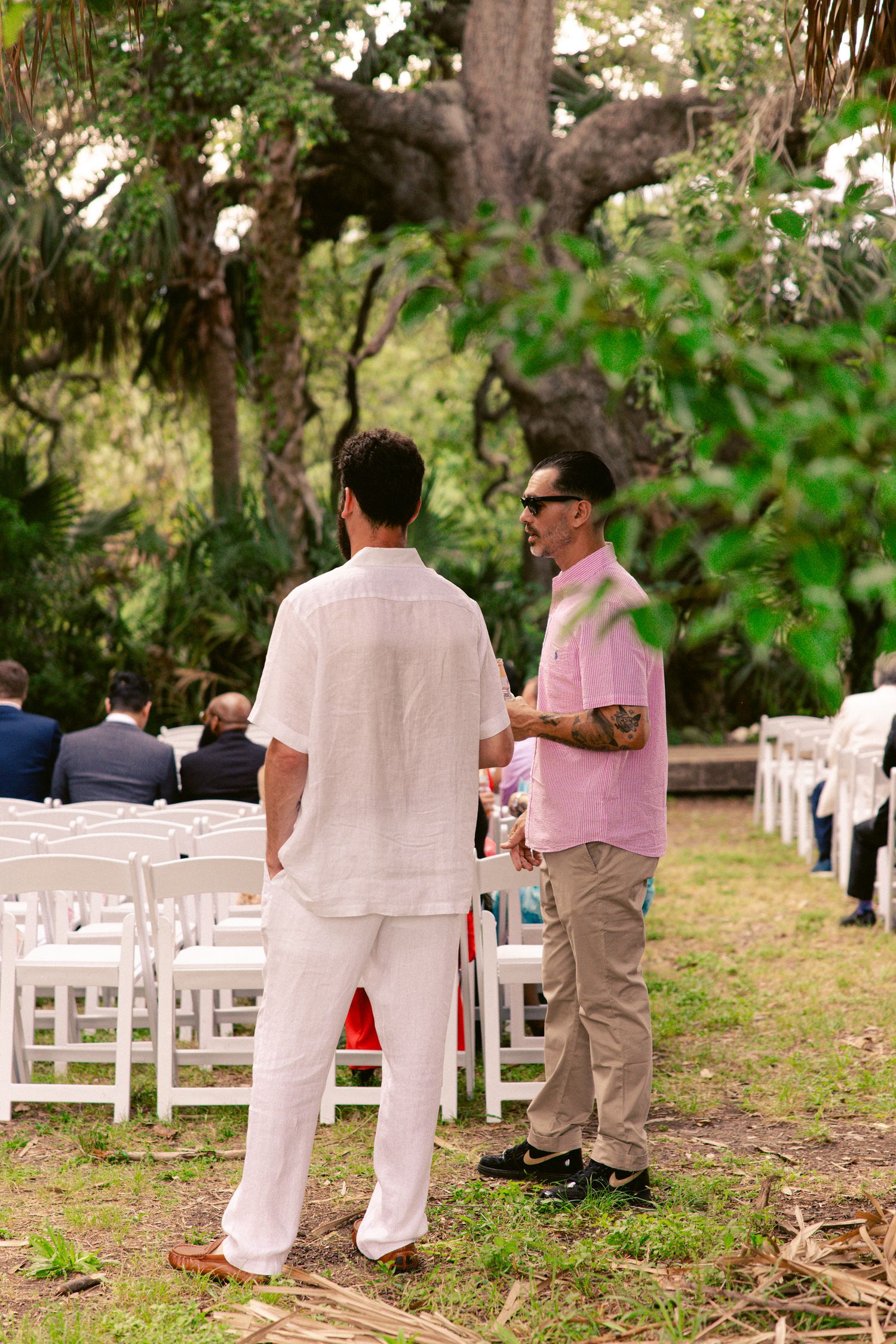 Two men in casual attire converse at an outdoor wedding ceremony with white chairs and lush greenery.