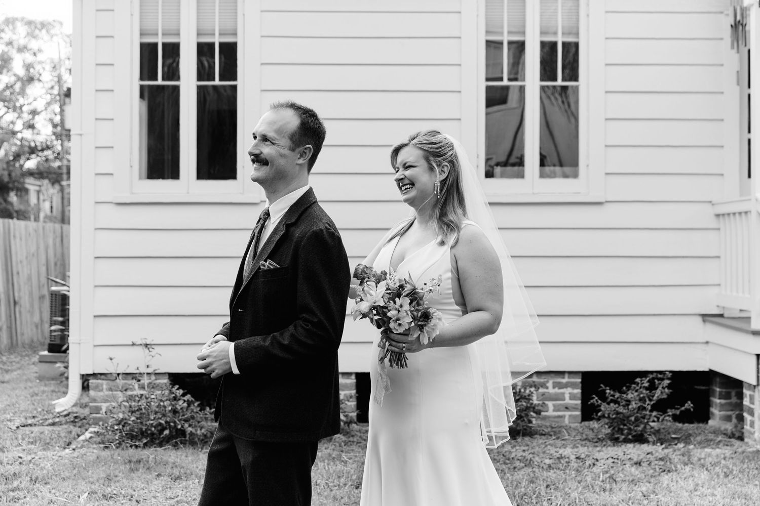 Black and white photo of bride and groom sharing a joyful moment outside a white building.