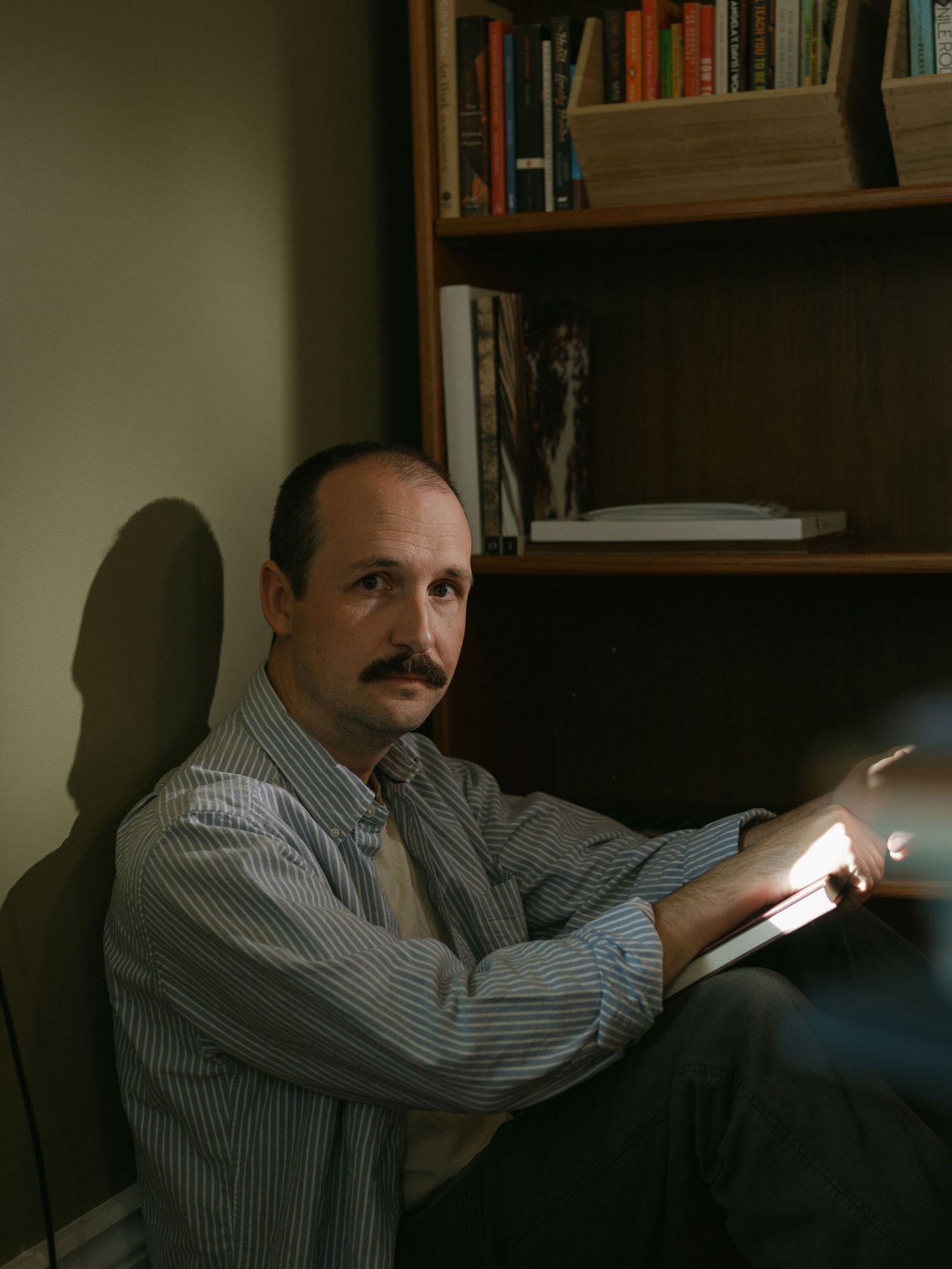 Man with mustache reading a glowing tablet beside a wooden bookshelf in dim light.
