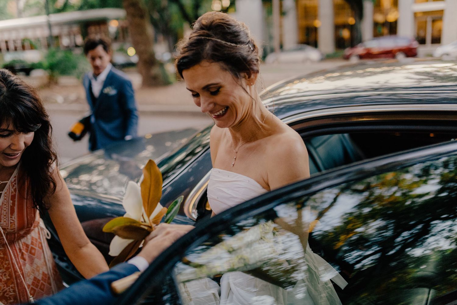 Radiant bride in strapless gown smiles while entering car assisted by bridesmaid after wedding.