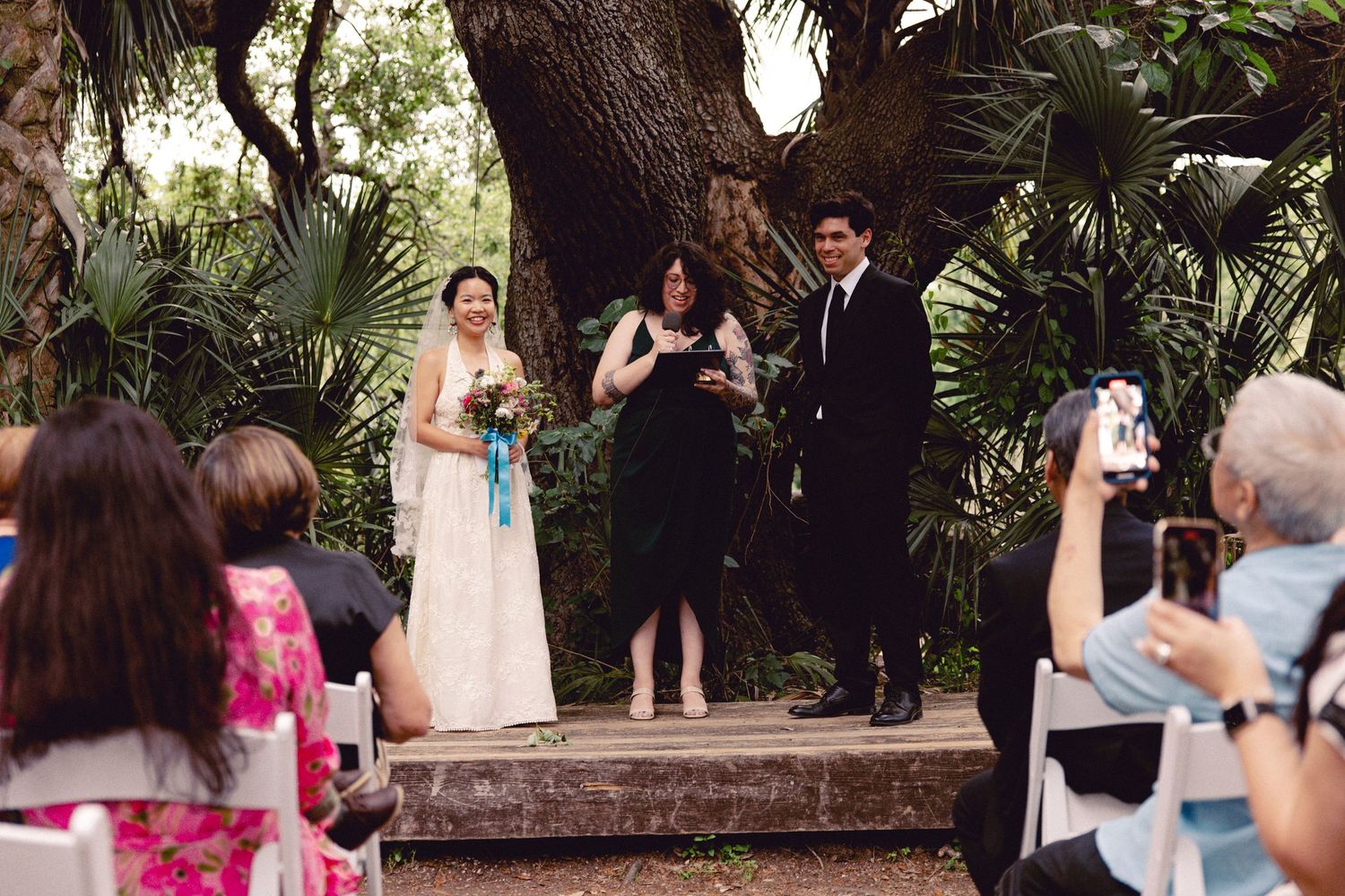 Bride in white lace gown and groom in black suit stand with officiant during outdoor garden wedding.