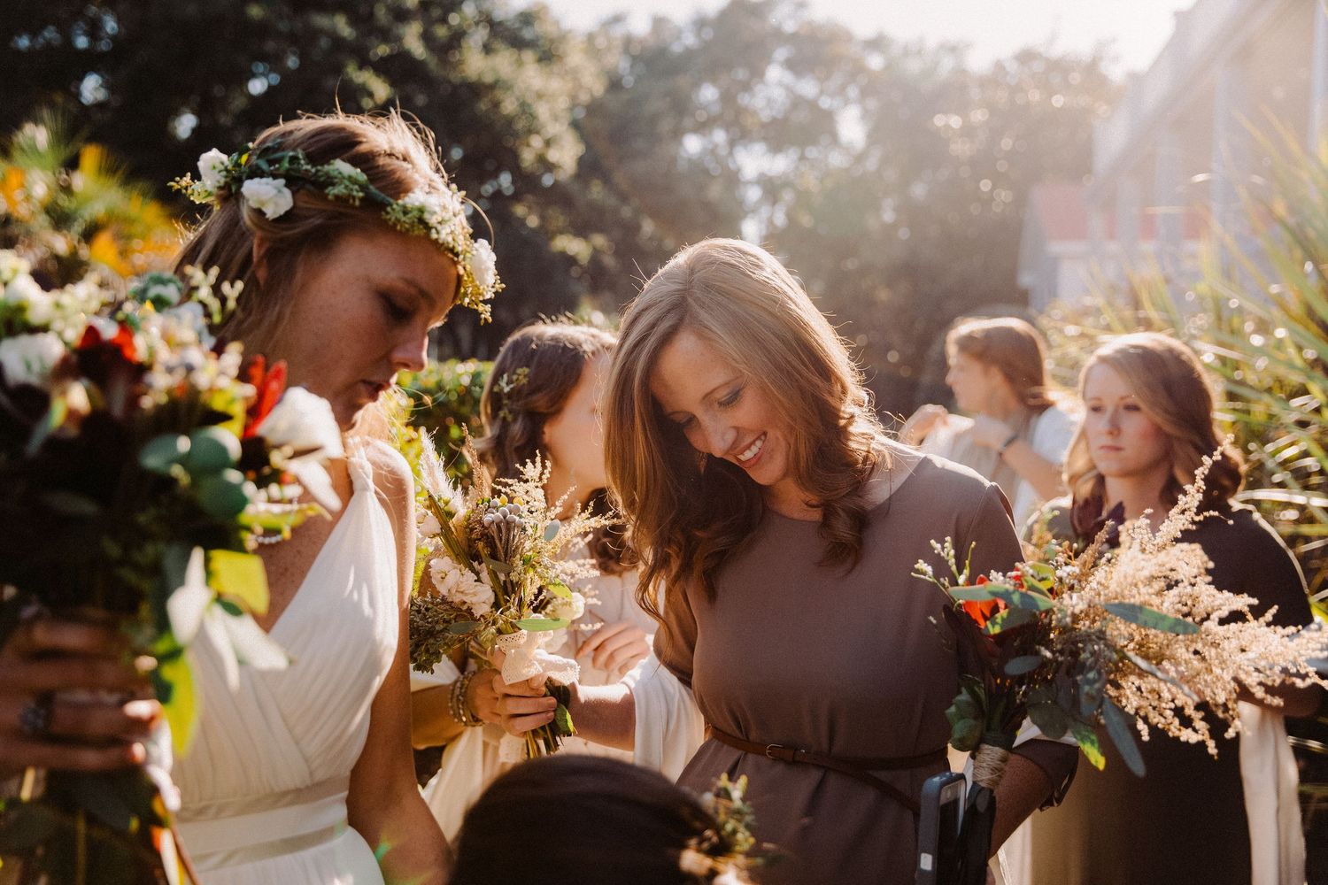 Bridesmaids holding wildflower bouquets in golden sunlight, one wearing a floral crown at an outdoor wedding.